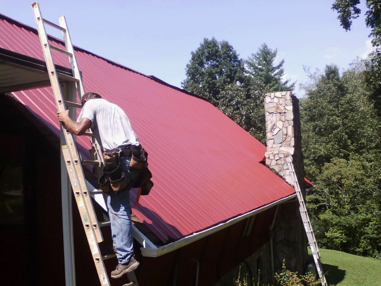 Roofer on a ladder beside a red metal roof and stone chimney — Soffit & Fascia Repair by Roofing Brothers in Satsop